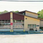 A beige fire station with a sloped roof, displaying a large American flag on a pole to the right. The station has glass garage doors and a sign that reads "Fire Station." The parking spaces in front of the building are marked with "No Parking" signs. Trees are in the background.