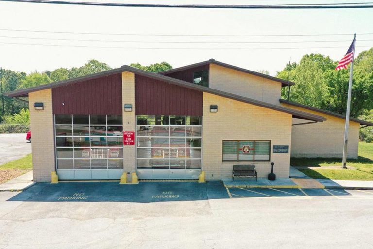 A beige fire station with a sloped roof, displaying a large American flag on a pole to the right. The station has glass garage doors and a sign that reads "Fire Station." The parking spaces in front of the building are marked with "No Parking" signs. Trees are in the background.