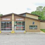 A modern fire station with a beige exterior and large glass doors labeled "RESCUE 82" and "ENGINE 42". Two yellow bollards are in front of the doors. The building is marked with the number "2" and has an American flag on a pole to the right. Trees are in the background.