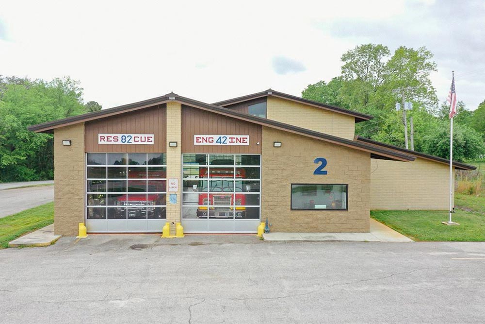 A modern fire station with a beige exterior and large glass doors labeled "RESCUE 82" and "ENGINE 42". Two yellow bollards are in front of the doors. The building is marked with the number "2" and has an American flag on a pole to the right. Trees are in the background.