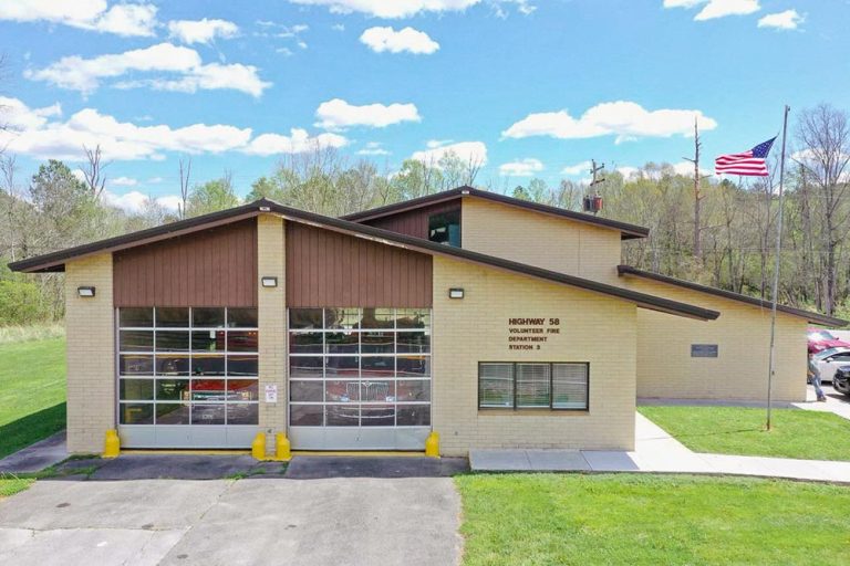 A modern fire station with a beige exterior and dark brown slanted roof situated in a lush green area. The station has large glass panel garage doors and an American flag flying on a pole to the right. Trees and a blue sky with clouds are in the background.