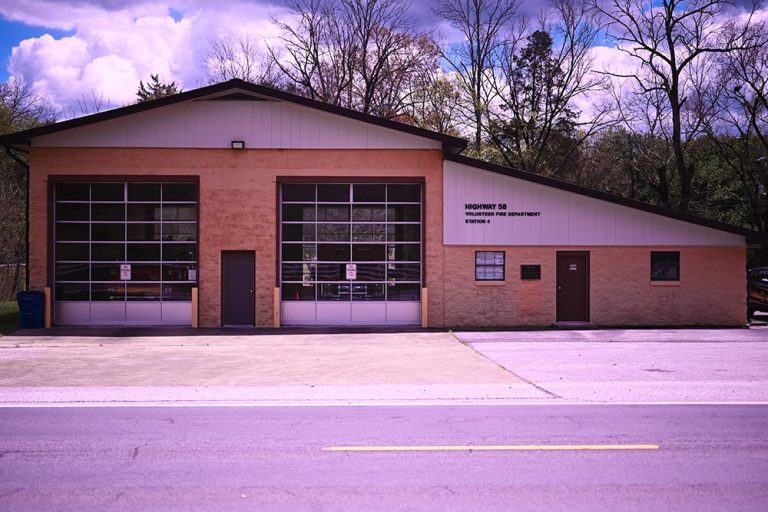 A one-story fire station with two large closed garage doors and a smaller side entrance. The building is labeled "Highway 50 Volunteer Fire Department Station 2." Surrounded by bare trees, the scene is lit by natural daylight under a partly cloudy sky.