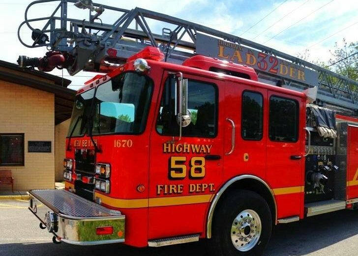 A red fire truck with the number 58 and "HIGHWAY FIRE DEPT" on the side is parked outside a beige brick building. The truck has a ladder extended above it, with "LAD32DER" visible on the ladder. Sunlight illuminates the scene, casting shadows.
