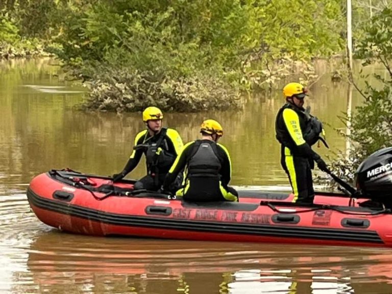 Three people in yellow helmets and black wetsuits ride an inflatable red and black rescue boat through a flooded area with trees and bushes partially submerged in the water.