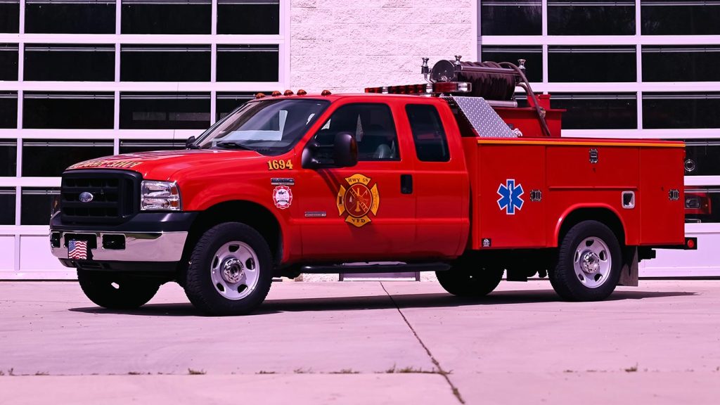 A red fire department utility truck is parked in front of a building with large glass-paned doors. The truck has various emergency symbols on its side, including a fire department emblem and a blue medical star of life symbol.