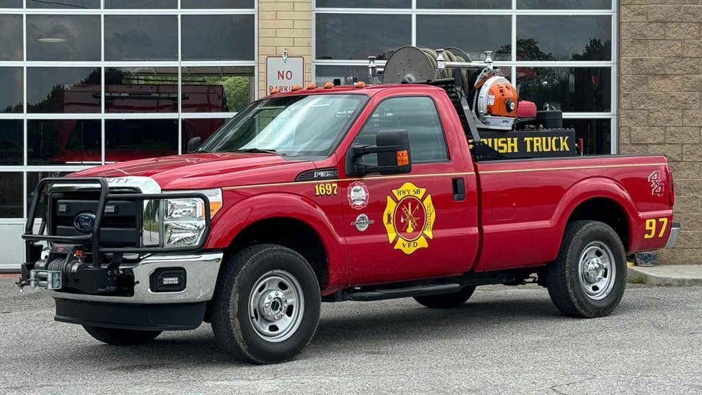 A red fire department brush truck, labeled "Brush Truck 97," is parked in front of a building with large garage doors. The vehicle has various firefighting equipment including hoses and a water tank mounted in the truck bed. The fire department logo is visible on the door.