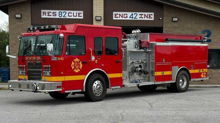 A red and yellow fire truck labeled "42" is parked outside a building with signs that read "RES CUE 82" and "ENG INE 42." The fire truck has various equipment compartments and a hose attached to its side.