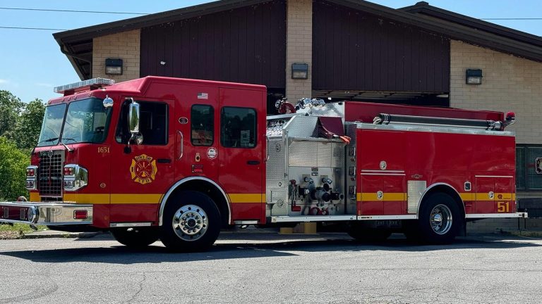 A bright red firetruck with yellow accents parked outside a brick fire station on a sunny day. The truck is equipped with hoses and firefighting equipment, and the number "51" is displayed on its side.