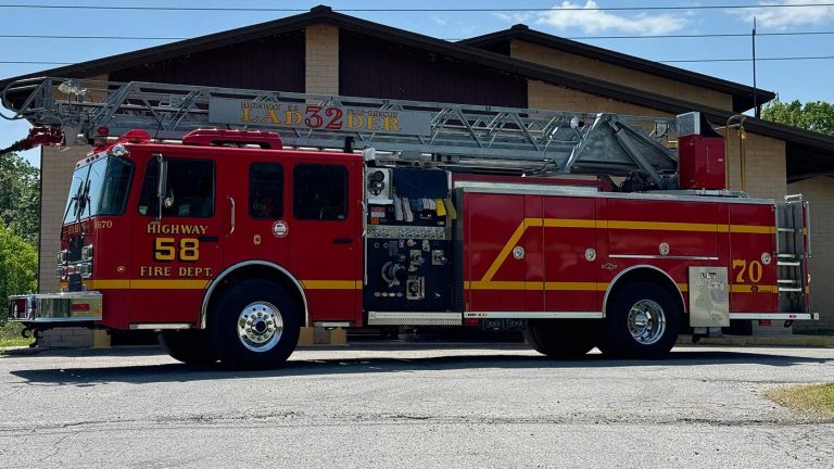 A red fire truck with the label "Highway Fire Dept. 58" and "Ladder 32" is parked outside a building. The truck features an extended ladder on top and various compartments, typical of a fire engine. The sky is clear and blue.