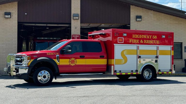 A bright red fire and rescue vehicle with "HIGHWAY 58 FIRE & RESCUE" written on the side is parked in front of a fire station. The vehicle has yellow stripes and emergency lights. The station building features a garage with open doors.