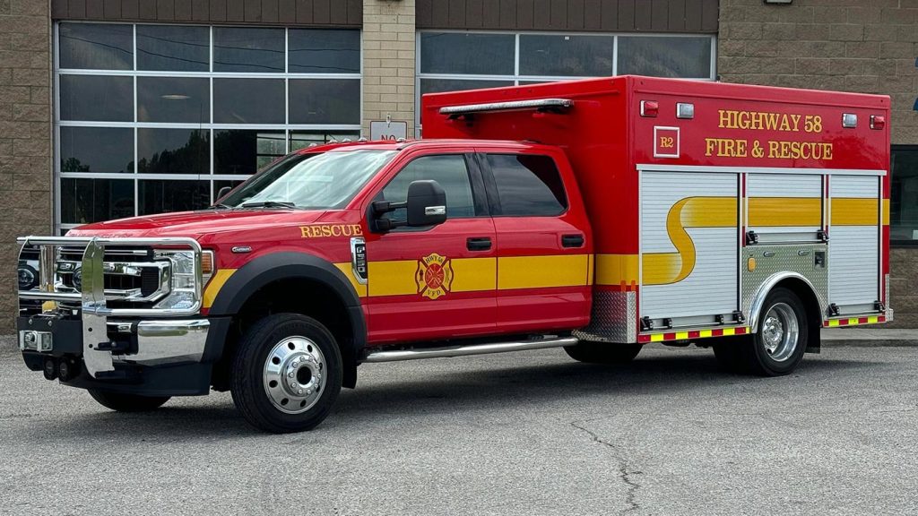 A red and yellow Highway 58 Fire & Rescue truck is parked outside a building. The truck features an extended cab and emergency lights on top, with the Rescue logo and large yellow stripe along the side. The background shows large windows and a brick wall.