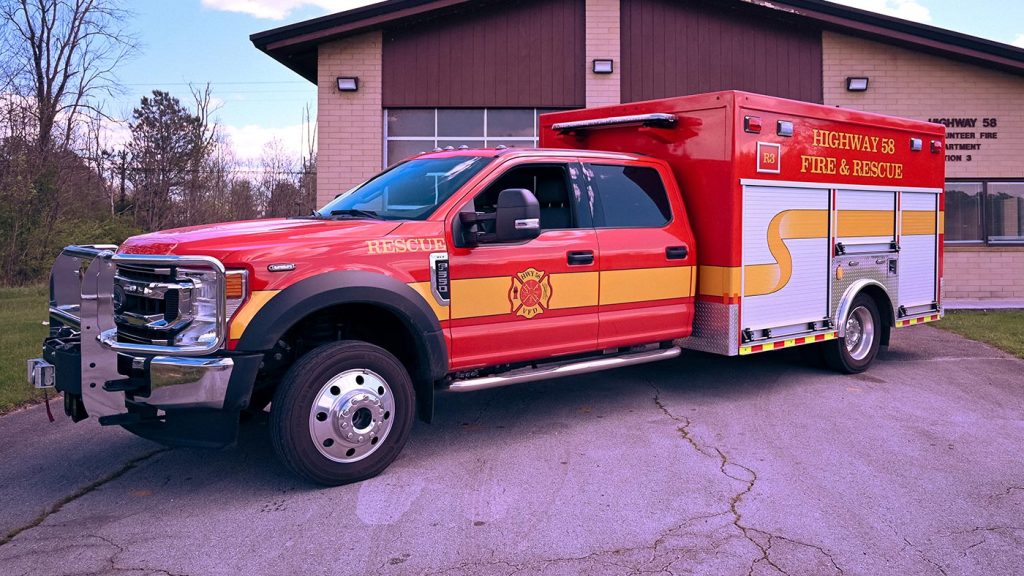 A bright red fire and rescue truck with yellow accents is parked outside a brick fire station. The truck is labeled "Highway 58 Fire & Rescue" and "Rescue" on its side. The fire station has a garage door and a glass window. Trees are visible in the background.