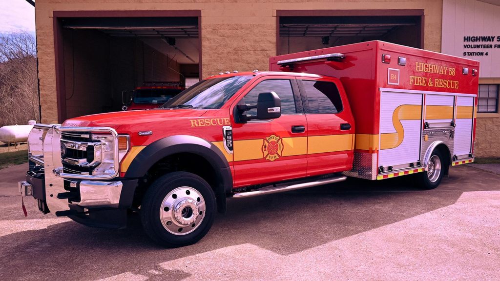A red and yellow fire and rescue truck is parked outside a fire station. The truck has a large cab and an extended rear section with various compartments. The text "HIGHWAY 58 FIRE & RESCUE" is visible on the side of the vehicle.