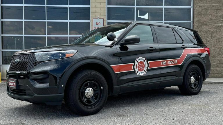 A black SUV with "Fire Rescue" and a fire department logo on the side is parked in front of a building with large windows and a "No Parking" sign. The vehicle has emergency lights installed on the front grille.