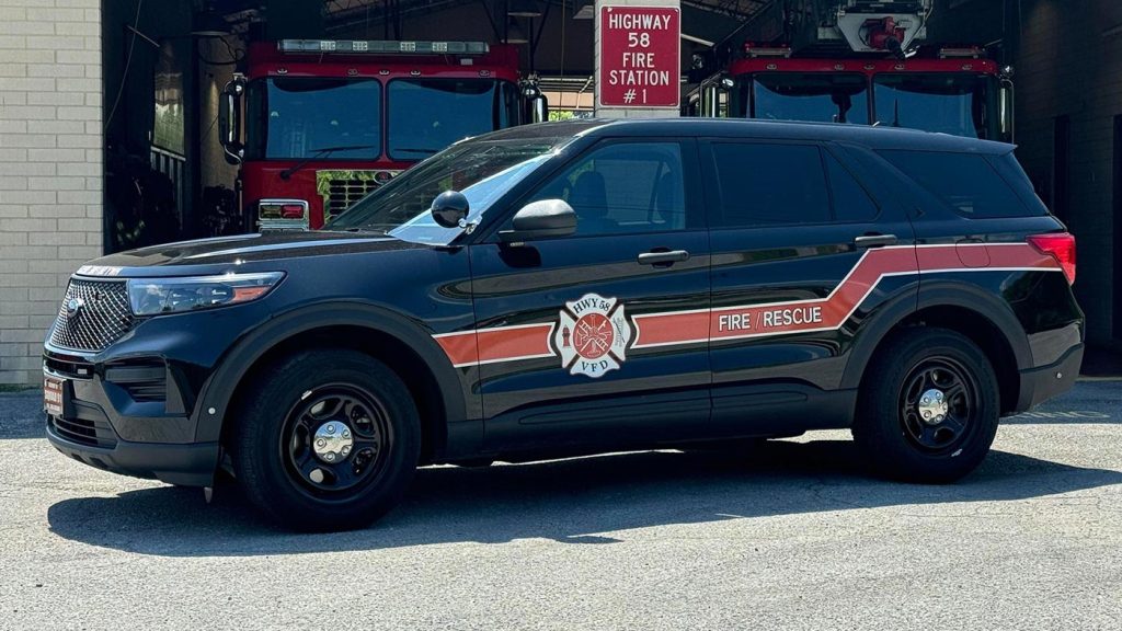 A black fire rescue SUV with red and white striping and the fire department emblem on the door is parked in front of a fire station. Two red fire trucks are visible in the background under the signage for "Highway 58 Fire Station #1.