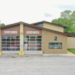 A fire station with two garage doors labeled "RESCUE 82" and "ENGINE 42". The station is number 2 and has an American flagpole to the right. A firetruck is visible inside through the glass doors. The building is surrounded by greenery and a paved driveway.