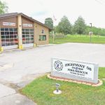 A fire station with a beige brick exterior and two large garage doors marked "RESCUE" and "4126." In front, a sign reads "Highway 58 Fire/Rescue Station 2." The surrounding area is grassy with trees, and an American flag is visible near the building.