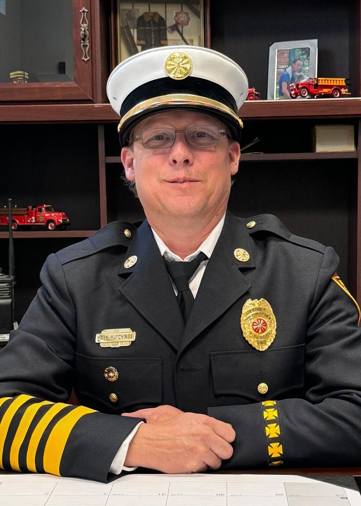 A fire chief dressed in a formal uniform sits at a desk. The chief is wearing a white hat, glasses, and a dark jacket with insignia and badges. Behind, there are shelves with small ornamental fire trucks and framed photos.