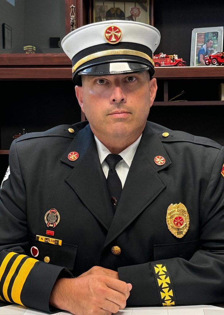 A firefighter in a formal uniform, including a white hat with a badge, sits at a desk. The uniform is adorned with various insignias and badges. Behind him, there are small model fire trucks and books on a wooden shelf.