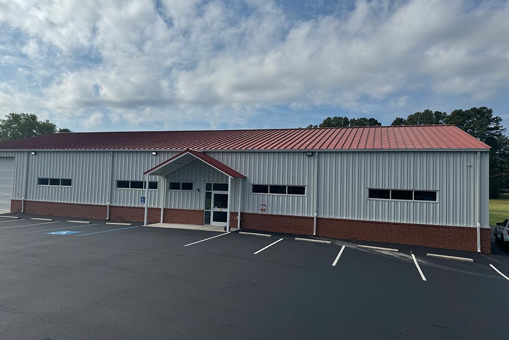 A modern single-story industrial building with a red metal roof and white siding, featuring multiple windows and a central glass entrance. The building sits on a large, freshly paved parking lot with marked spaces, including a disabled spot near the entrance.