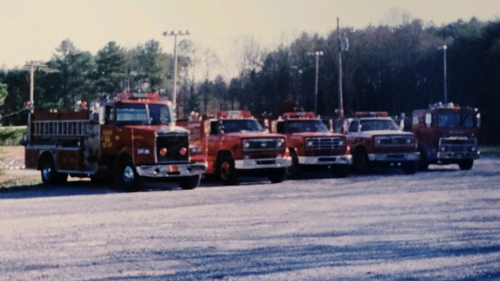 A row of red fire trucks is parked on a gravel lot, surrounded by leafless trees in the background. The trucks are of varying sizes, with some featuring ladders and lights. The scene appears to be a rural or suburban area.