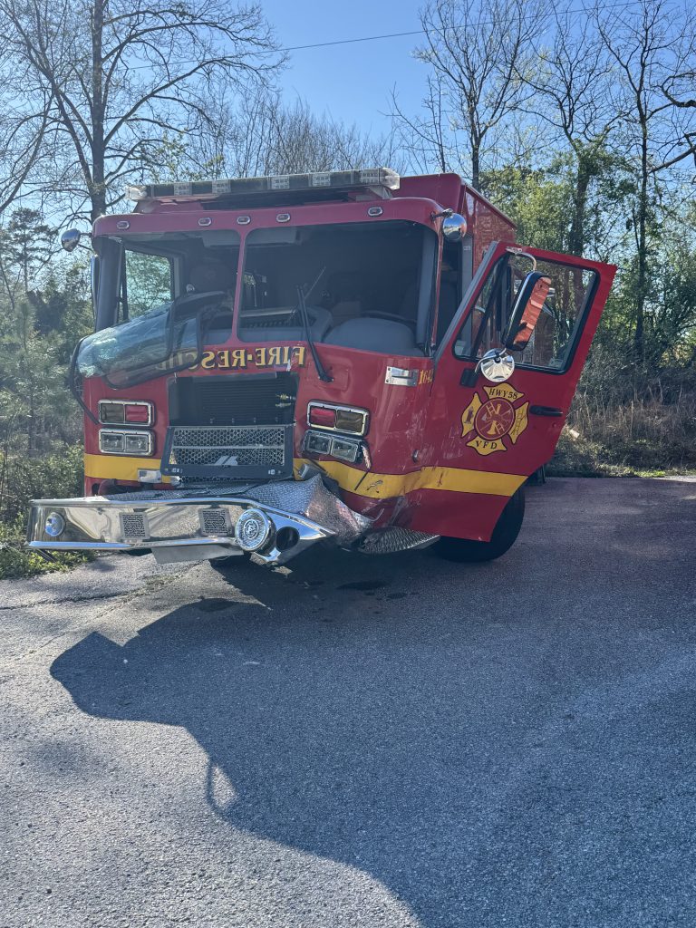 A red fire truck with visible front-end damage is parked on a paved area. Its front bumper is bent, and the passenger door is open. Trees and grass are in the background under a clear blue sky.