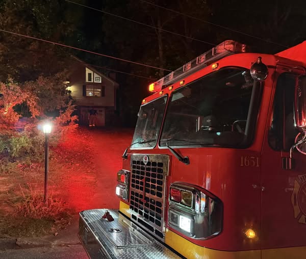 A red fire truck with flashing lights is parked at night outside a house on a tree-lined street, illuminating the area with red light.