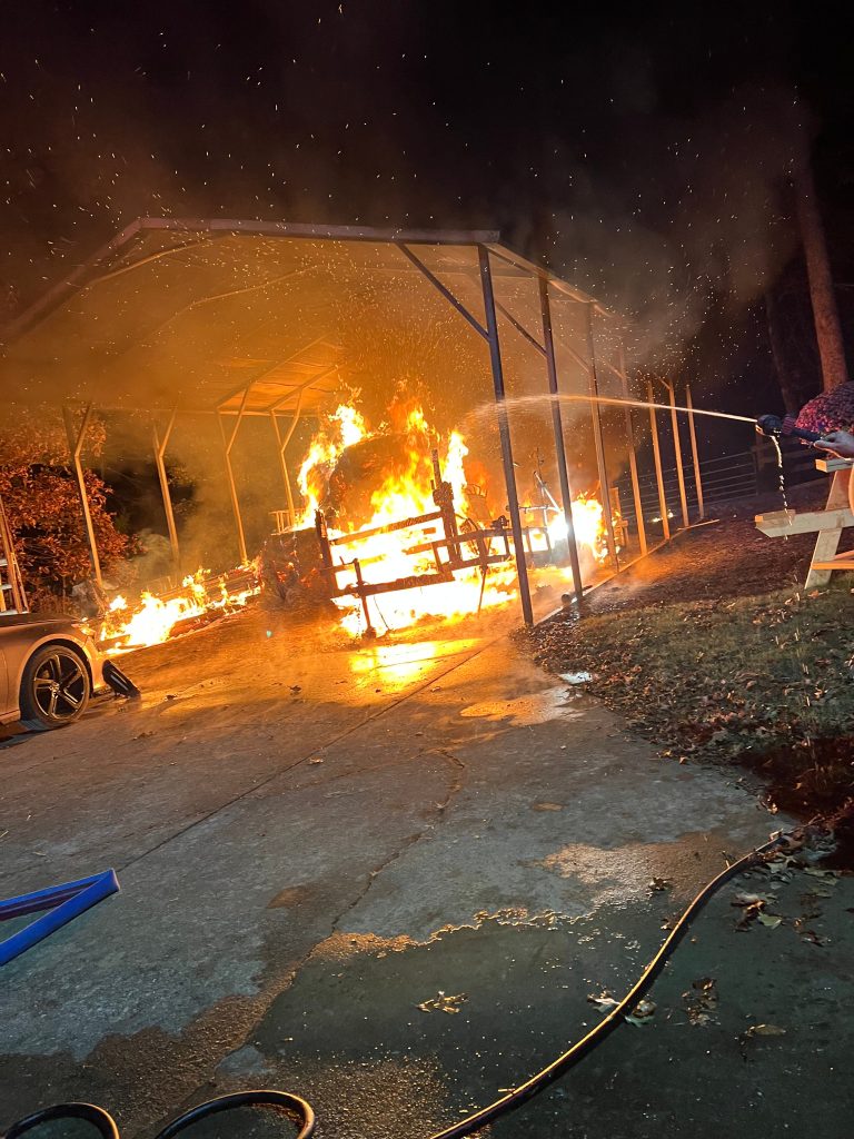 A large fire burns under a metal carport at night as a person sprays water from a hose to extinguish the flames. Nearby, a car and a wooden picnic table are visible. Orange flames and smoke light up the dark surroundings.