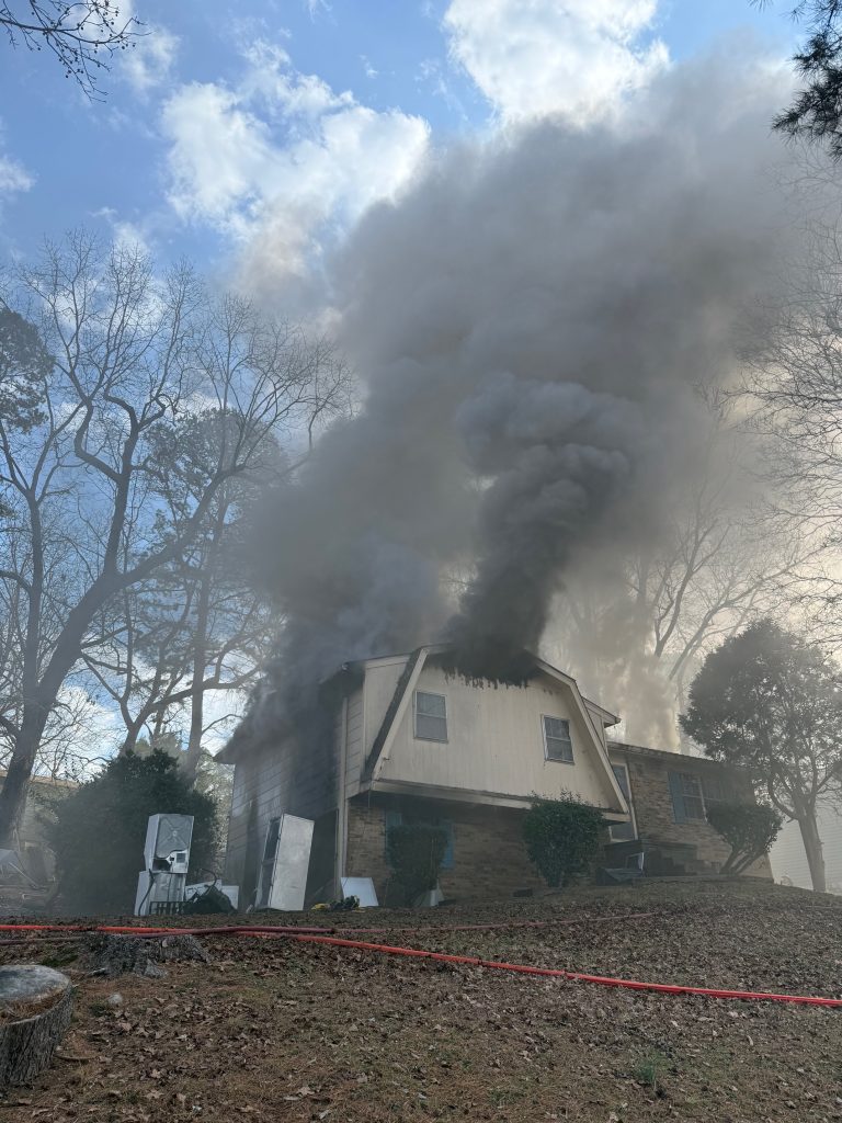 A large house emits thick black smoke from its upper floor windows as a fire burns inside; trees surround the house and the sky is partly cloudy.