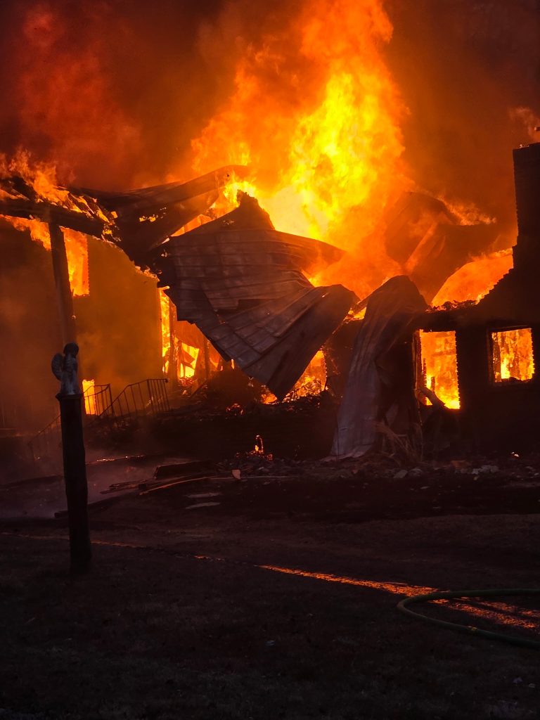 A raging fire engulfs a building at night, with large flames and thick smoke consuming the structure. Debris is collapsing and glowing orange, while the ground in front is dark and partially lit by the fire.