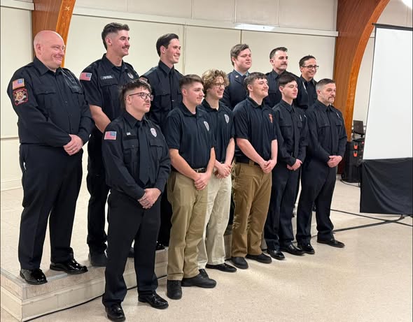 A group of twelve people, mostly in dark uniforms, pose together indoors for a formal photo. Eleven stand in two rows while one stands slightly elevated on a step. A large projection screen is visible in the background.