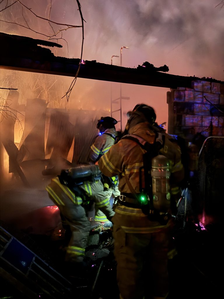 Two firefighters in full gear work amid smoke and flames inside a burning structure at night, surrounded by debris and illuminated by emergency lights.