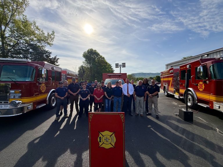 A group of firefighters and community members stand in sunlight between two fire trucks, posing for a photo outdoors near a podium with a fire department emblem. Trees and a blue sky with clouds are visible in the background.